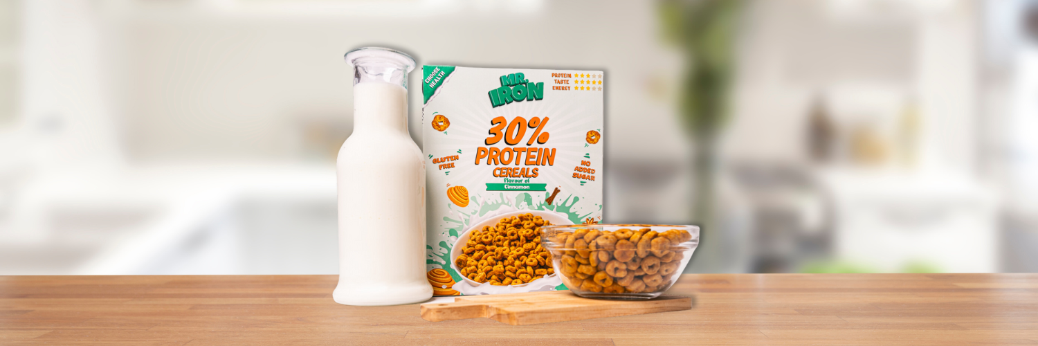 Cutie de cereale proteice Mr. Iron cu 30% proteine si aroma de scortisoara, fara gluten sau zahar adaugat, alaturi de o sticla de lapte si un bol plin, pe un blat de bucatarie.